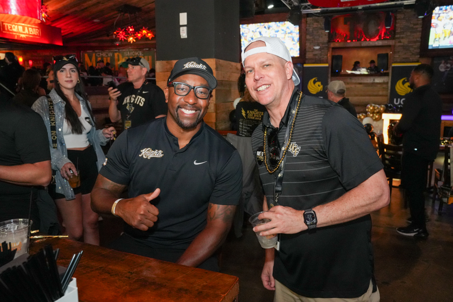 Two men at a crowded bar pose for a photo; one in a black athletic shirt and cap gives a thumbs-up, the other in a black striped polo with a white cap holds a drink.