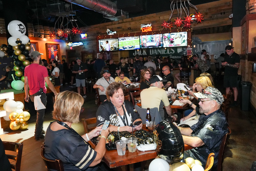 A busy sports bar with groups of people seated at wooden tables, festive gold and white balloons, and multiple TV screens above a bar area showing games or programs.
