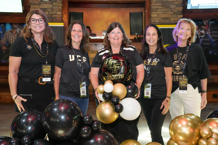 Five women in UCF-themed shirts pose and smile for a photo, holding a black, gold, and white balloon bouquet at an indoor event.