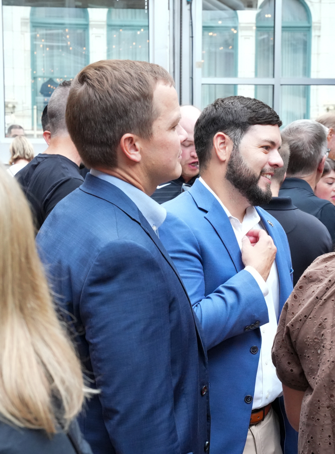 Two men in blue blazers stand among a crowd, smiling, as they chat at an outdoor event outside glass doors.