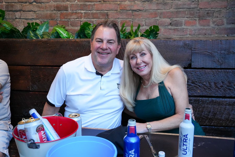 A smiling couple sits at a table with drinks and a brick wall behind them, posing for a photo at a social gathering.