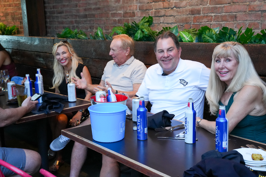 Group of smiling adults seated at a restaurant table with drinks and a blue ice bucket, enjoying a casual meal.