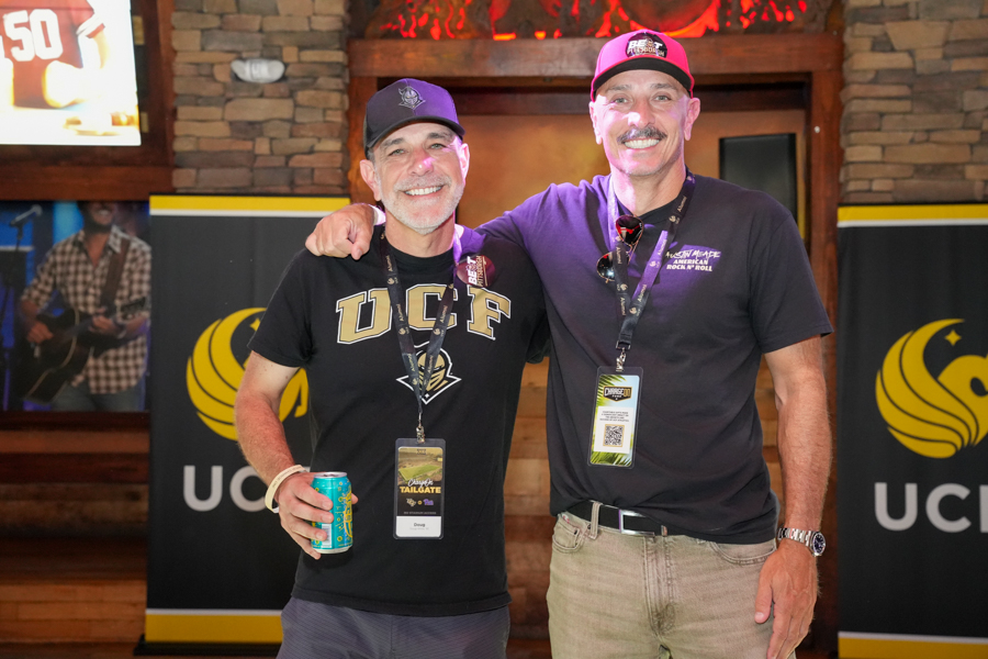 Two smiling men with event lanyards pose with arms around each other at a tailgate-style event, one in a UCF shirt holding a can of drink.