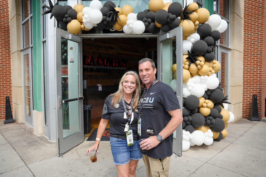 Smiling couple poses at the entrance of a venue framed by a black, white, and gold balloon arch, each wearing event badges.