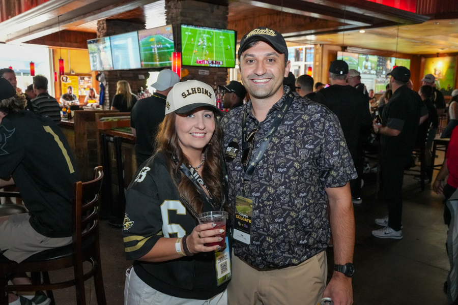 Two smiling fans pose together at a busy sports bar, the woman in a football jersey and cap holding a red drink while the man stands beside her with a lanyard and cap in front of several TV screens showing games.