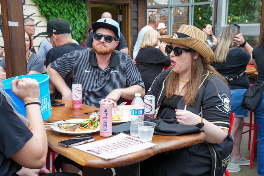 Group of people dining at an outdoor patio, two in the foreground wearing hats and sunglasses, with plates and drinks on a wooden table.