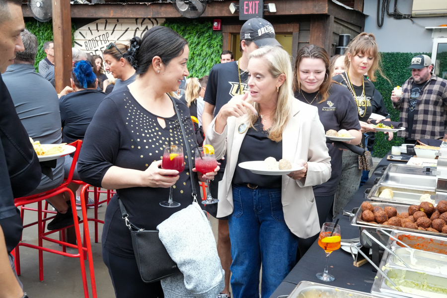Two women chat while holding plates and red cocktails at a crowded outdoor buffet event.