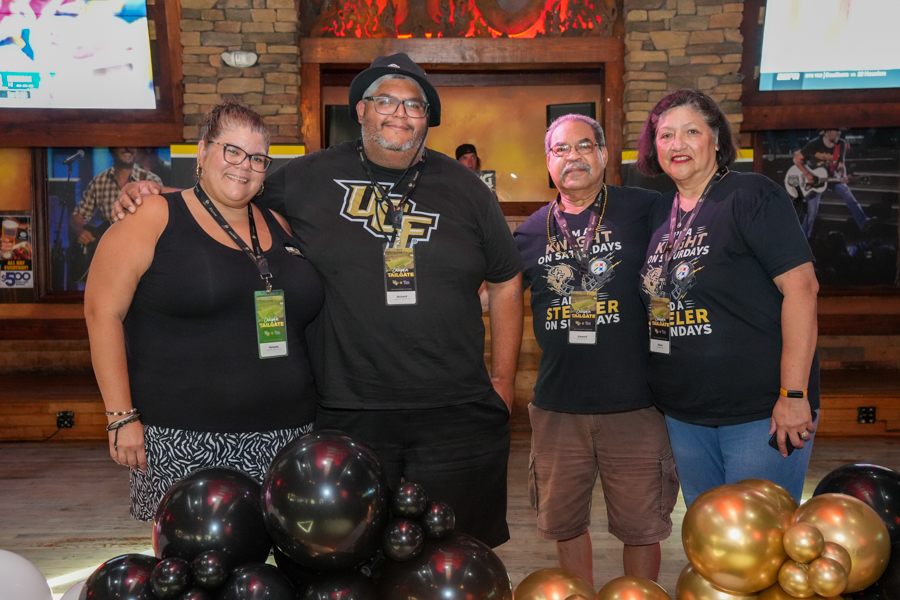 Group of four adults posing together at a venue, wearing event lanyards and black shirts, with black and gold balloons in front.