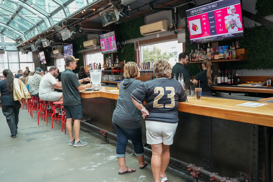 People stand and sit at a long wooden bar inside a bright, glass-ceiling venue, watching sports screens and ordering drinks.