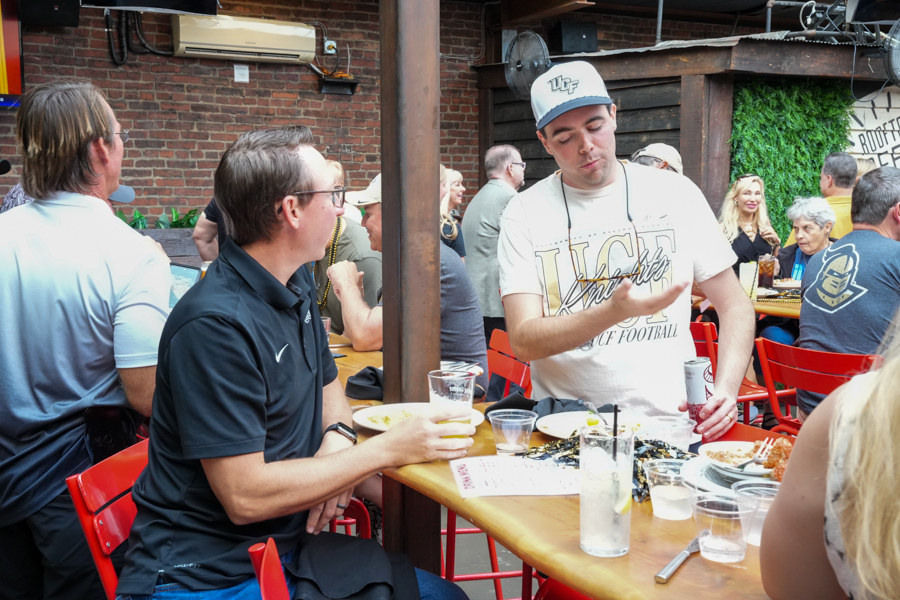 Two men sit at a wooden table outdoors, chatting over drinks as others dine around them at a casual bar or patio.