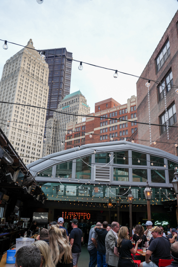Crowded outdoor bar on a city rooftop under string lights and a glass pavilion, with tall skyscrapers in the background.