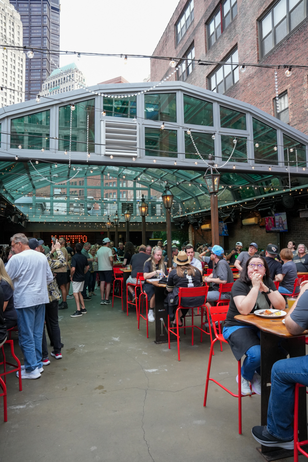 Crowded outdoor dining area under a glass canopy with string lights and bright red stools, in a city courtyard.