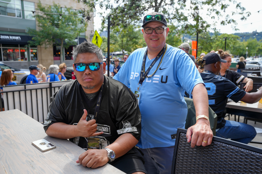 Two men at an outdoor event posing for the camera; one seated in a black jersey with blue mirrored sunglasses, the other standing in a light blue shirt with a cap and lanyard, resting a hand on a chair.