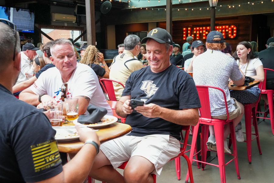 Group of friends at a busy bar/restaurant terrace, seated on red metal stools and chatting over drinks while one man texts on his phone in the foreground.