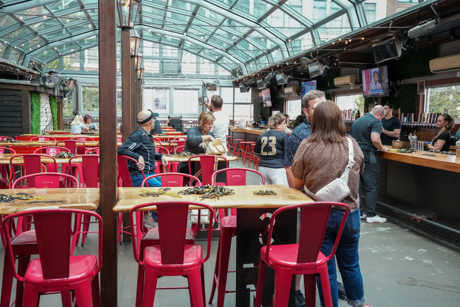 Glass-roofed beer hall with red metal chairs and wooden tables; patrons order drinks at a long bar on the right.