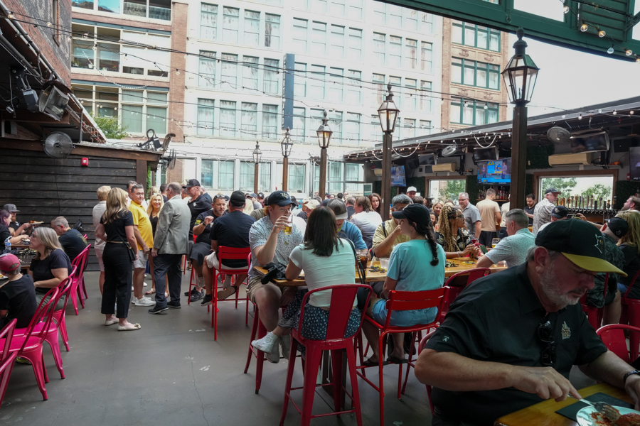 Crowded outdoor urban patio with people seated at red chairs around wooden tables, eating and chatting under string lights.