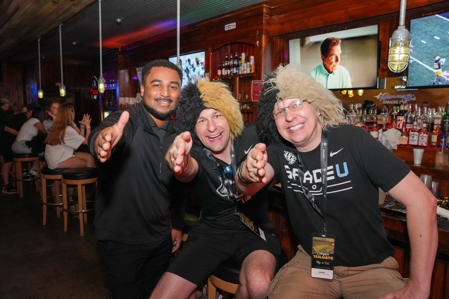 Three men at a bar pose for a photo, two wearing furry hats, all reaching toward the camera with smiles behind the counter and bottles in view.