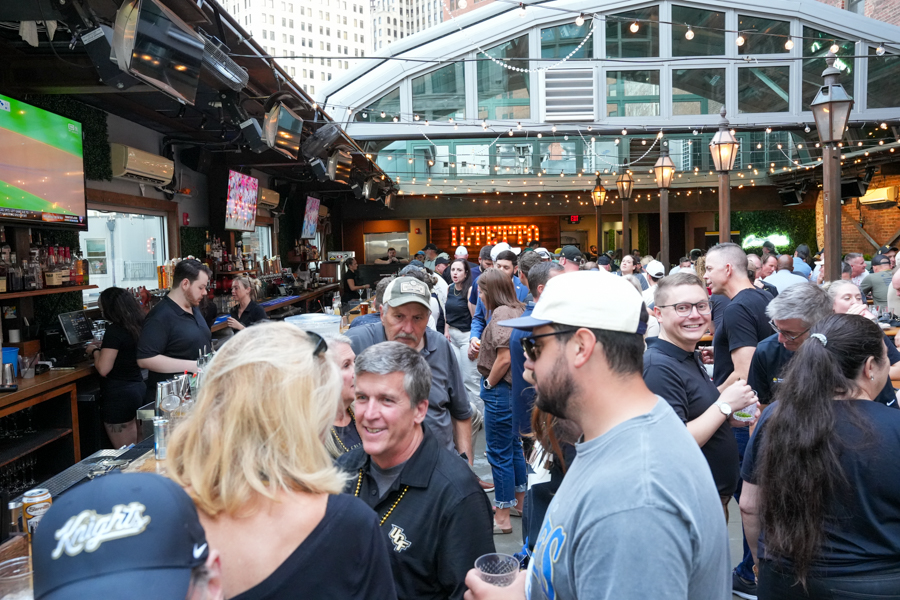 Crowded rooftop bar with string lights, people chatting and holding drinks near a long wooden bar.