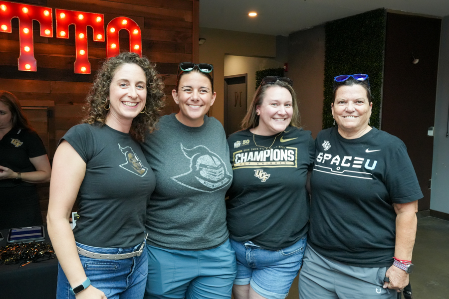 Four women pose together indoors, smiling at the camera, wearing dark T-shirts and jeans.