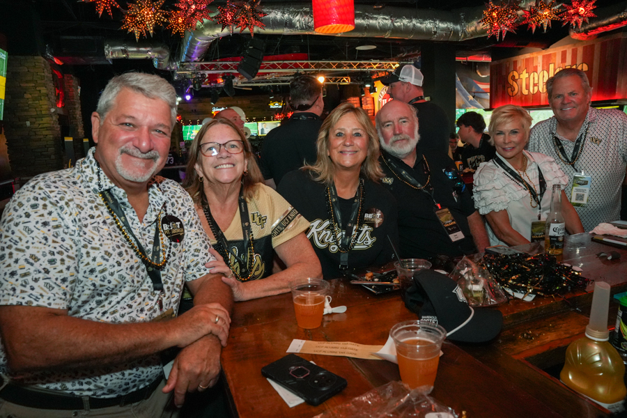 Group of six adults at a bar, smiling for the camera; festive decorations and drinks on the counter behind them.
