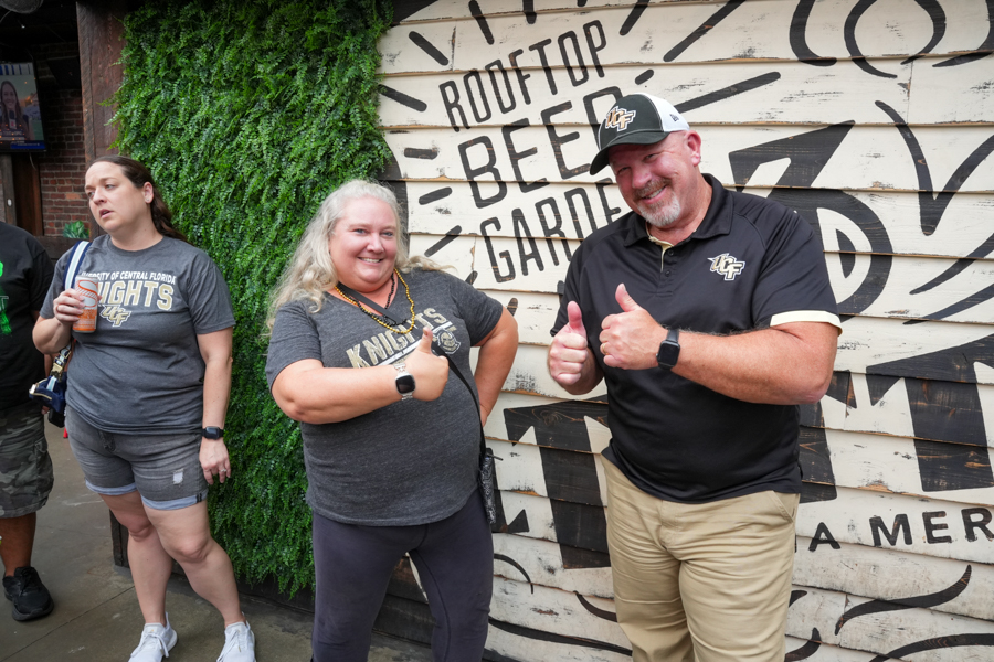 Three adults pose and thumbs‑up in front of a Rooftop Beer Garden mural.