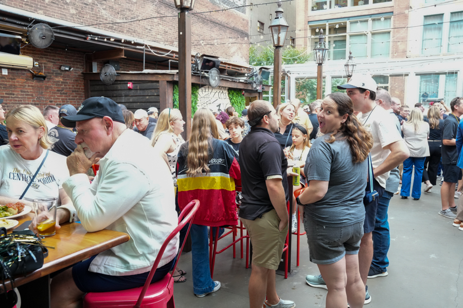 Crowd of people socializing at an outdoor courtyard, with a long wooden table and red chairs near a brick wall.
