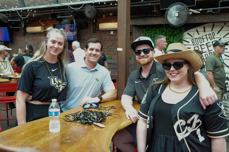 Four friends sit at a wooden table in a casual outdoor bar, smiling at the camera with drinks and beads on the table behind them
