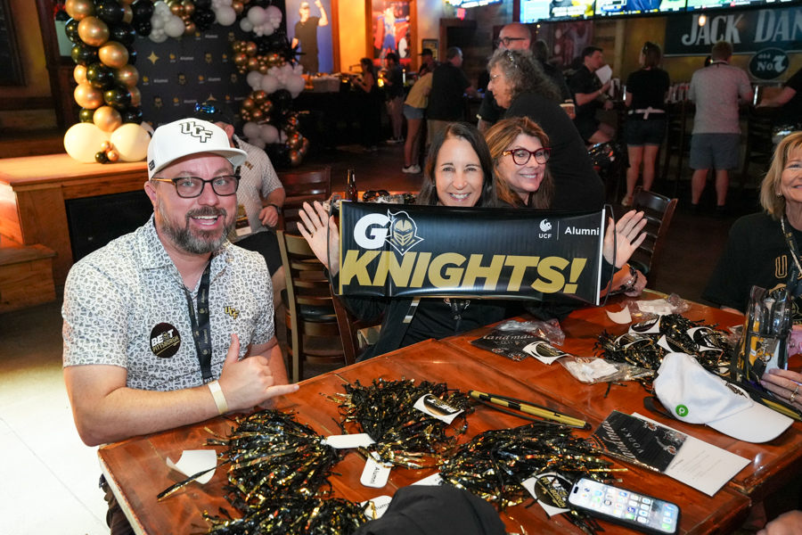 Group of smiling alumni seated at a table with a bold "GO KNIGHTS!" banner, celebrating at a UCF event with decorations and others in the background.