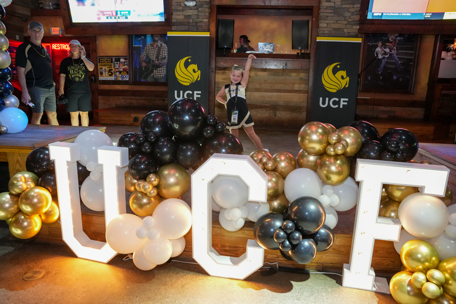 A young girl in a black-and-white outfit poses with one arm raised behind oversized illuminated letters spelling UCF, surrounded by gold, white, and black balloons at a venue stage.
