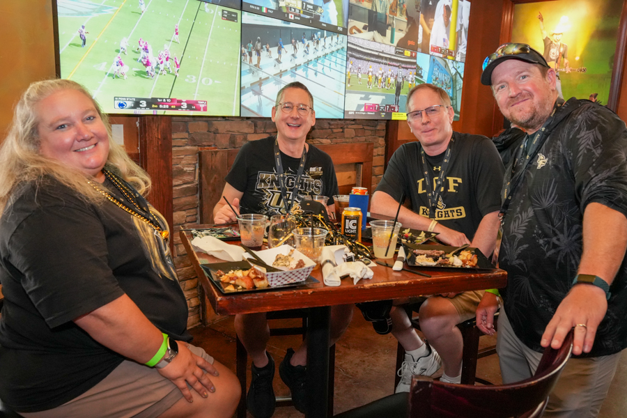 Four adults seated at a wooden table in a sports bar, smiling at the camera; plates of food and drinks on the table, with multiple TV screens showing sports in the background.