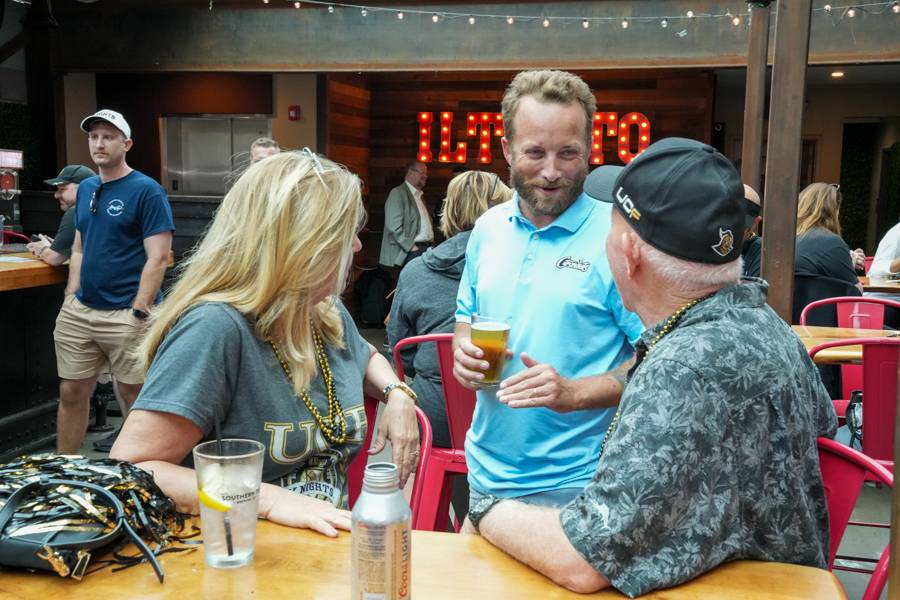 Three adults chatting at an outdoor bar table with drinks, string lights overhead and a red neon sign in the background.