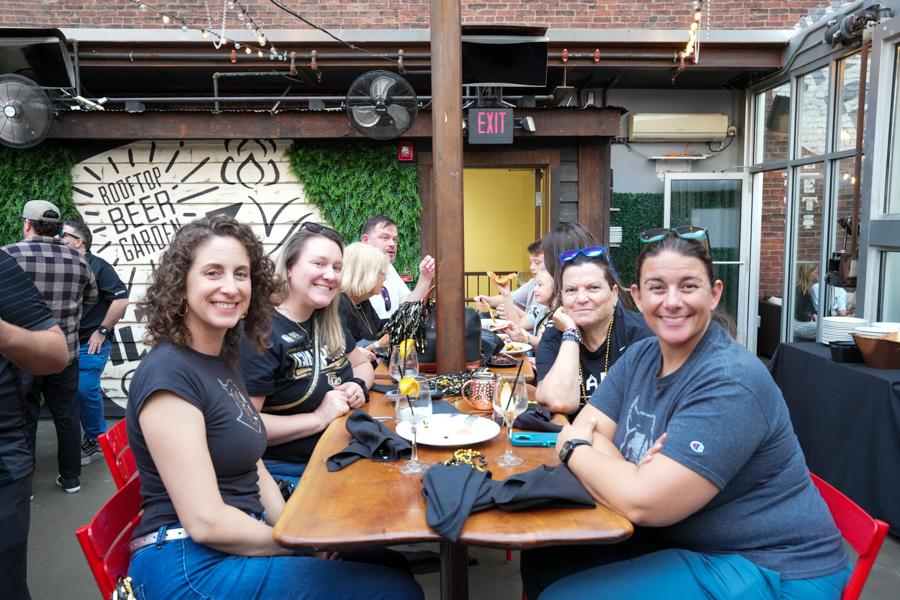 Group of friends seated along a long wooden table at a rooftop beer garden, smiling and chatting with drinks and plates in front of them.