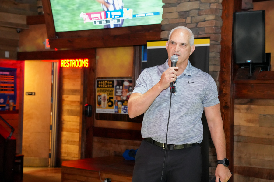 Man speaking into a handheld microphone at an indoor venue with wood panels and a neon restrooms sign in the background.