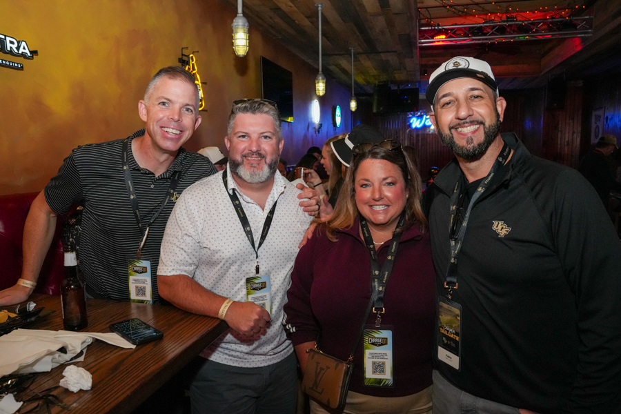 Four friends posing together at a bar, wearing event lanyards and smiling at the camera.