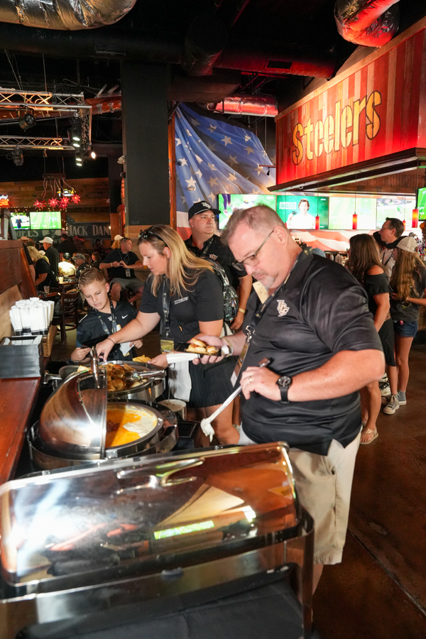 People line up at a buffet in a sports bar with a Steelers sign in the background, ready to eat.