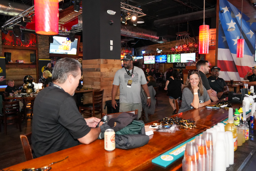 Bar scene with patrons at a wooden counter, red pendant lights, and a large American flag mural in the background.