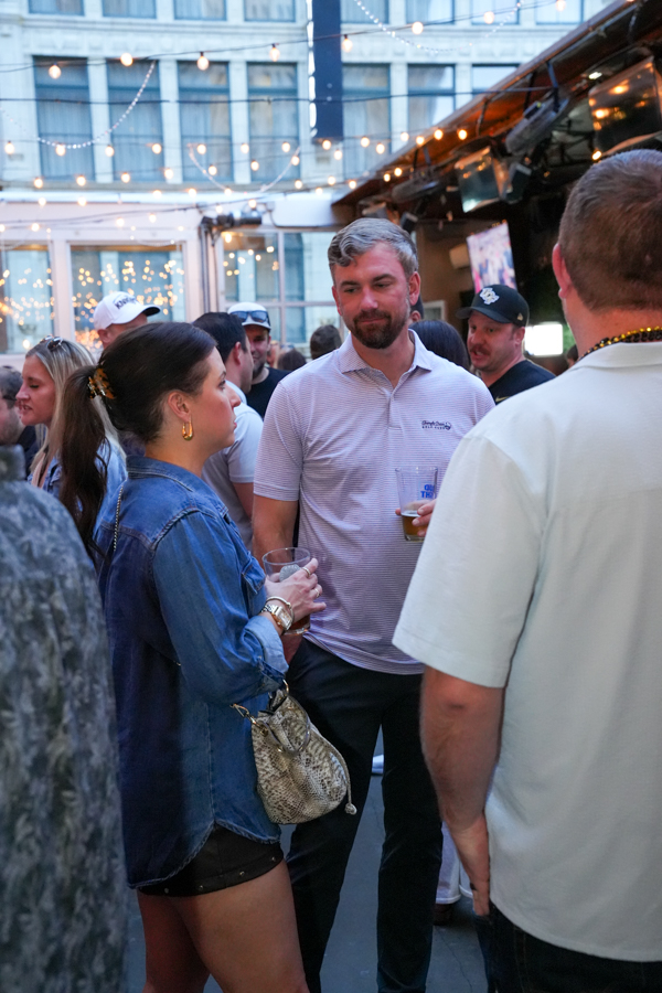 People mingling at an outdoor evening party with string lights; a man with a beer talks to a woman in a denim jacket.