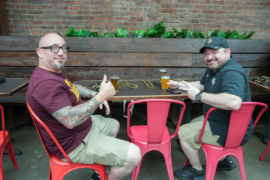 Two men sit at an outdoor wooden table, each holding a glass of beer and smiling at the camera.