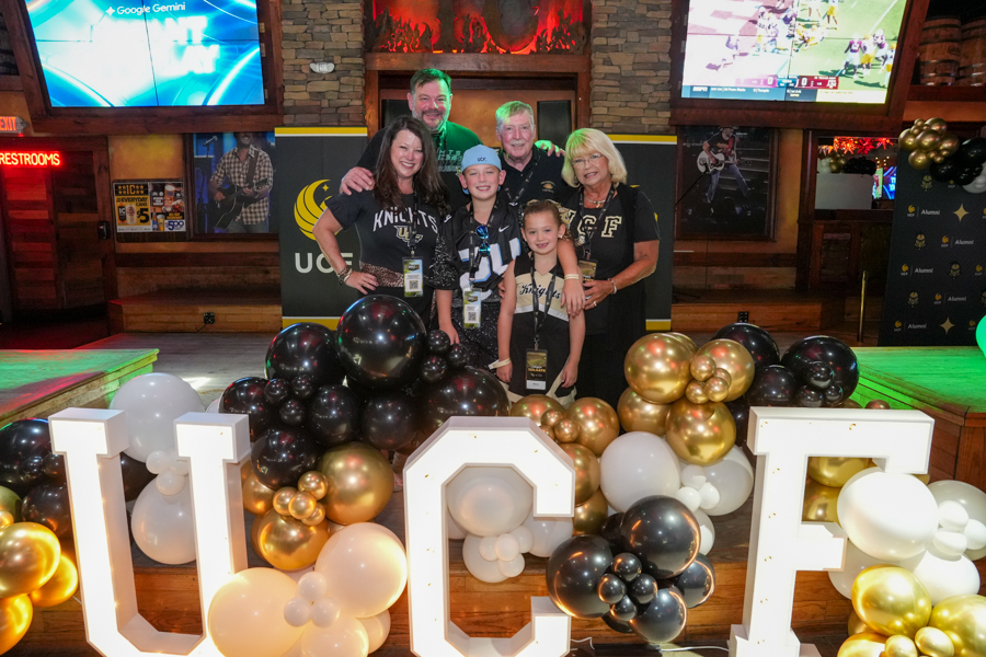 Group of six people posing for a photo behind large white illuminated letters surrounded by black, white, and gold balloons at a UCF-themed event in a bar.