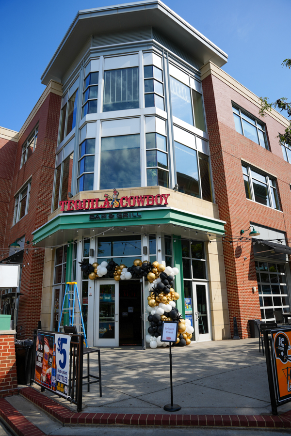 Exterior of Tequila Cowboy Bar & Grill storefront with teal awning and a black, white, and gold balloon arch at the entrance, celebrating a launch or event.