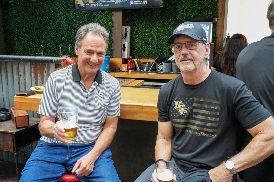Two men sit at a wooden bar smiling at the camera, each holding a beer.