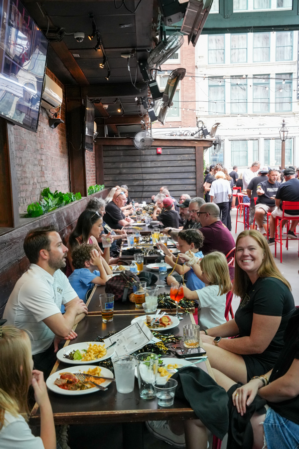 Large group dining at a long communal table in a brick-walled restaurant, eating and chatting with plates of food and drinks visible.