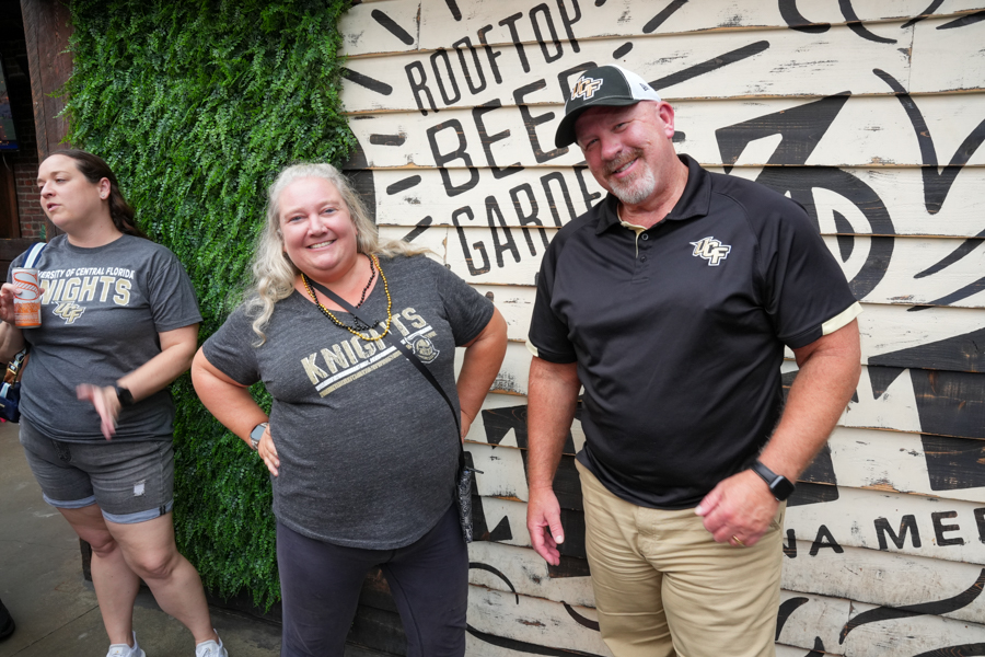 Three adults posing in front of a large mural with black graphic designs; center woman wears a gray Knights shirt, man on right in a black polo, all smiling.