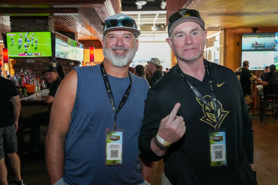 Two smiling men posing at a bar or event venue, wearing lanyards with badges; one in a blue sleeveless shirt, the other in a black long-sleeve shirt with a logo, the man on the right showing a raised finger gesture.