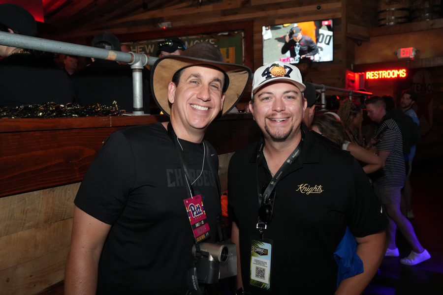 Two men smiling for a photo at a rustic venue, one with a wide-brim hat and camera around his neck, the other in a cap and polo with a badge nearby.