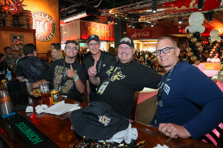 Four men at a festive bar posing for the camera; one gives a thumbs-up, background shows a Steelers sign and balloons.