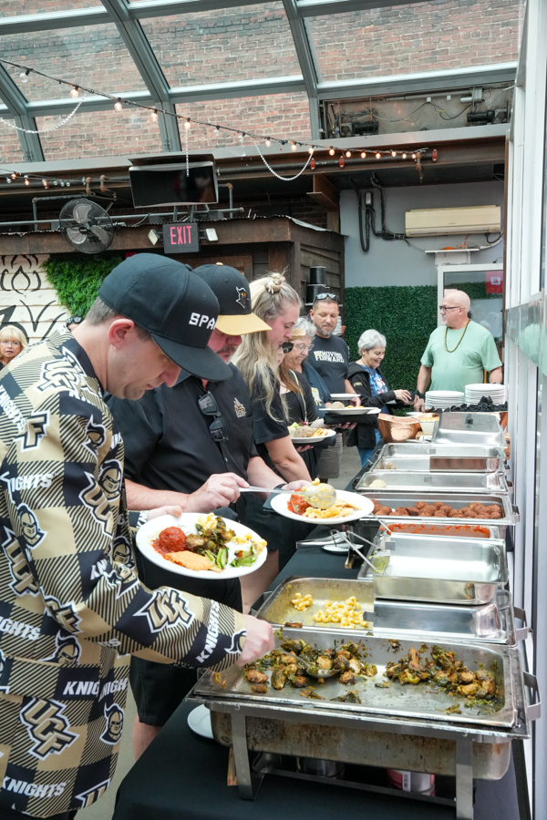 People in a casual buffet line serving themselves from chafing dishes outdoors under a glass roof, plates piled with food.