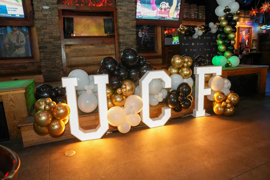 Large illuminated letters spelling 'UCF' surrounded by gold, black, and white balloons at a festive venue backdrop