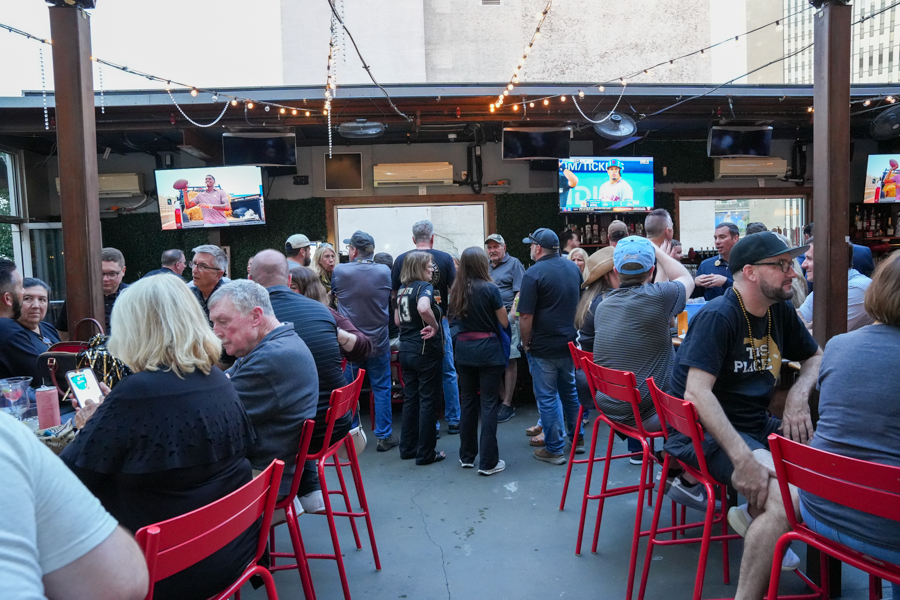 Crowded outdoor bar patio with red chairs, string lights, and multiple TVs showing sports as patrons mingle.