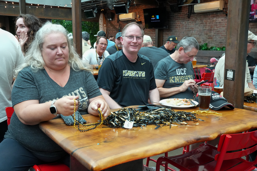 Two women and a man sit at a wooden bar table, sorting gold beads while others eat and chat in a casual outdoor-style setting with Knights gear nearby.